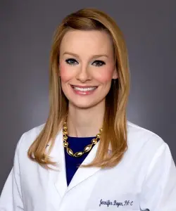A woman with straight blonde hair wearing a white medical coat and blue top against a dark backdrop.