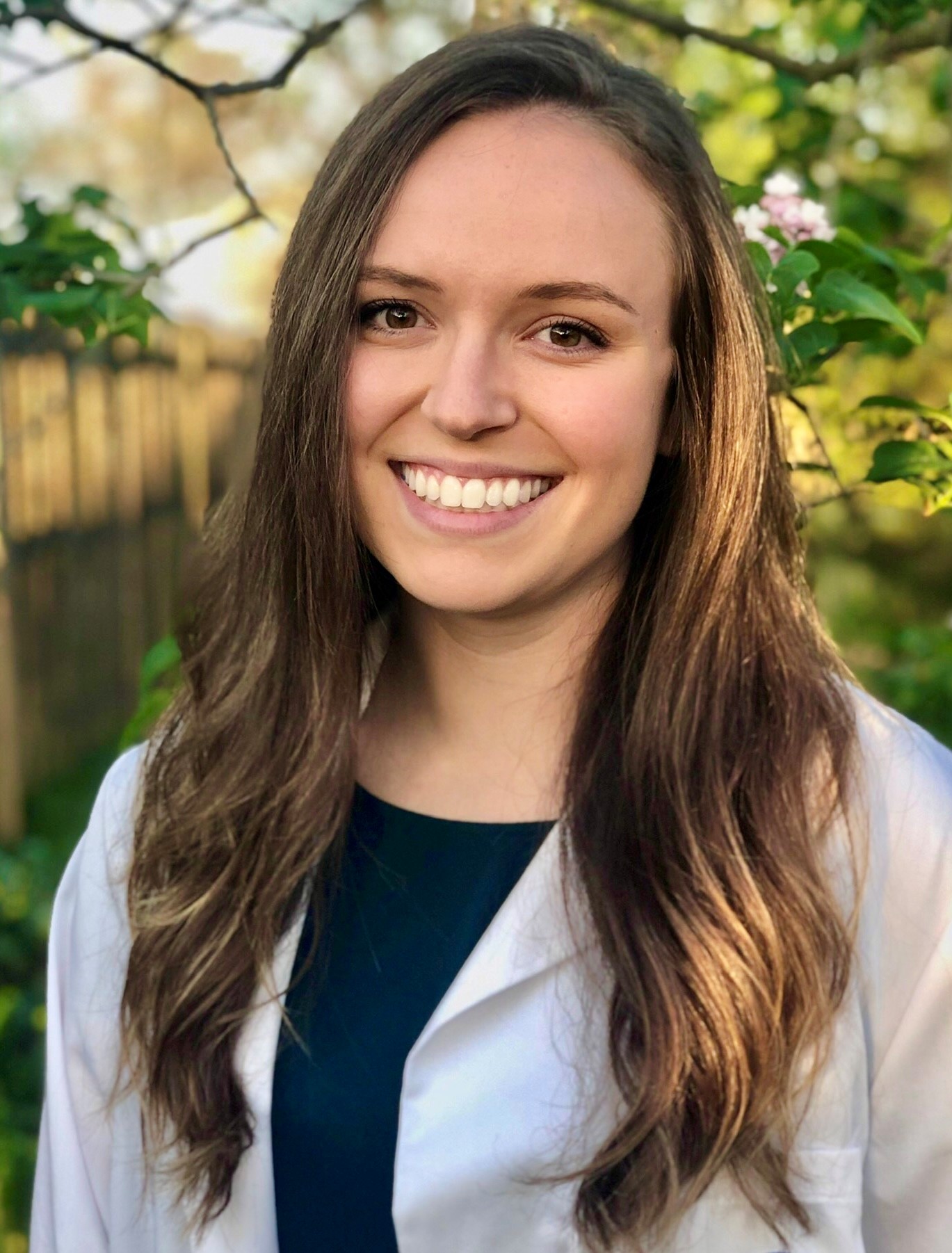 A woman with long brown hair wearing a white lab coat and dark top smiles with greenery behind her.