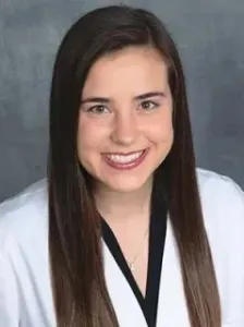 A woman with long brown hair, wearing a white coat over a black top against a gray background.