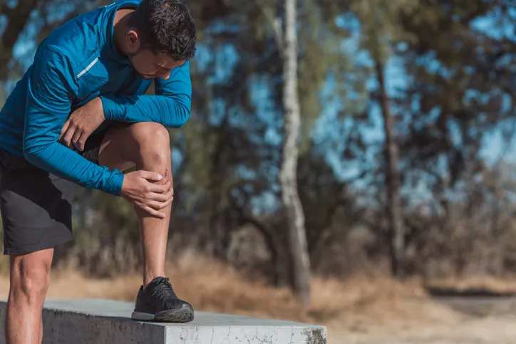 A man in athletic clothing stands outdoors, leaning on one knee while holding his leg.