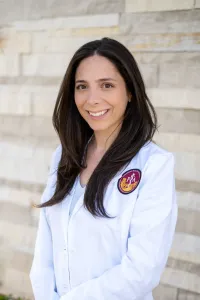 A woman with long dark hair wearing a white lab coat stands in front of a light-colored brick wall.