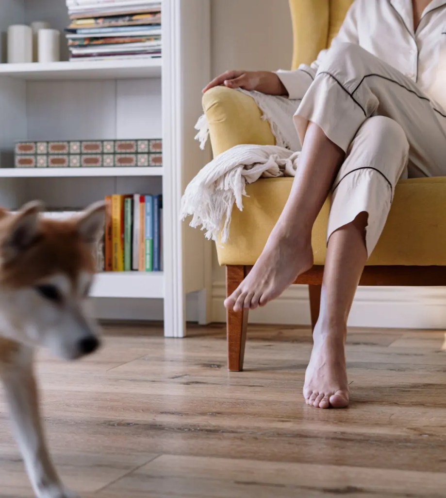 Person sitting barefoot in a yellow armchair with a blanket and a dog walking past.