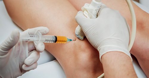 A medical professional injects medicine into a patient's knee using a syringe, with an ultrasound.